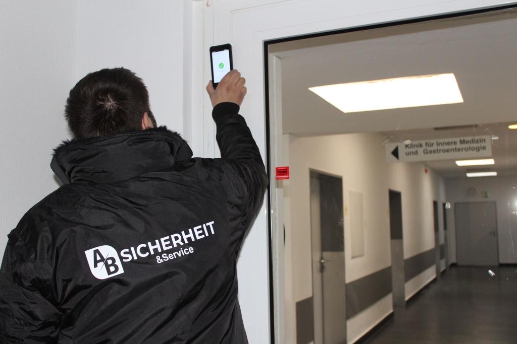 Ein Mann mit Jacke mit dem Aufdruck absicherheit.de steht mit dem Rücken zum Betrachter und hält ein Telefon für einen Scan an eine Tür